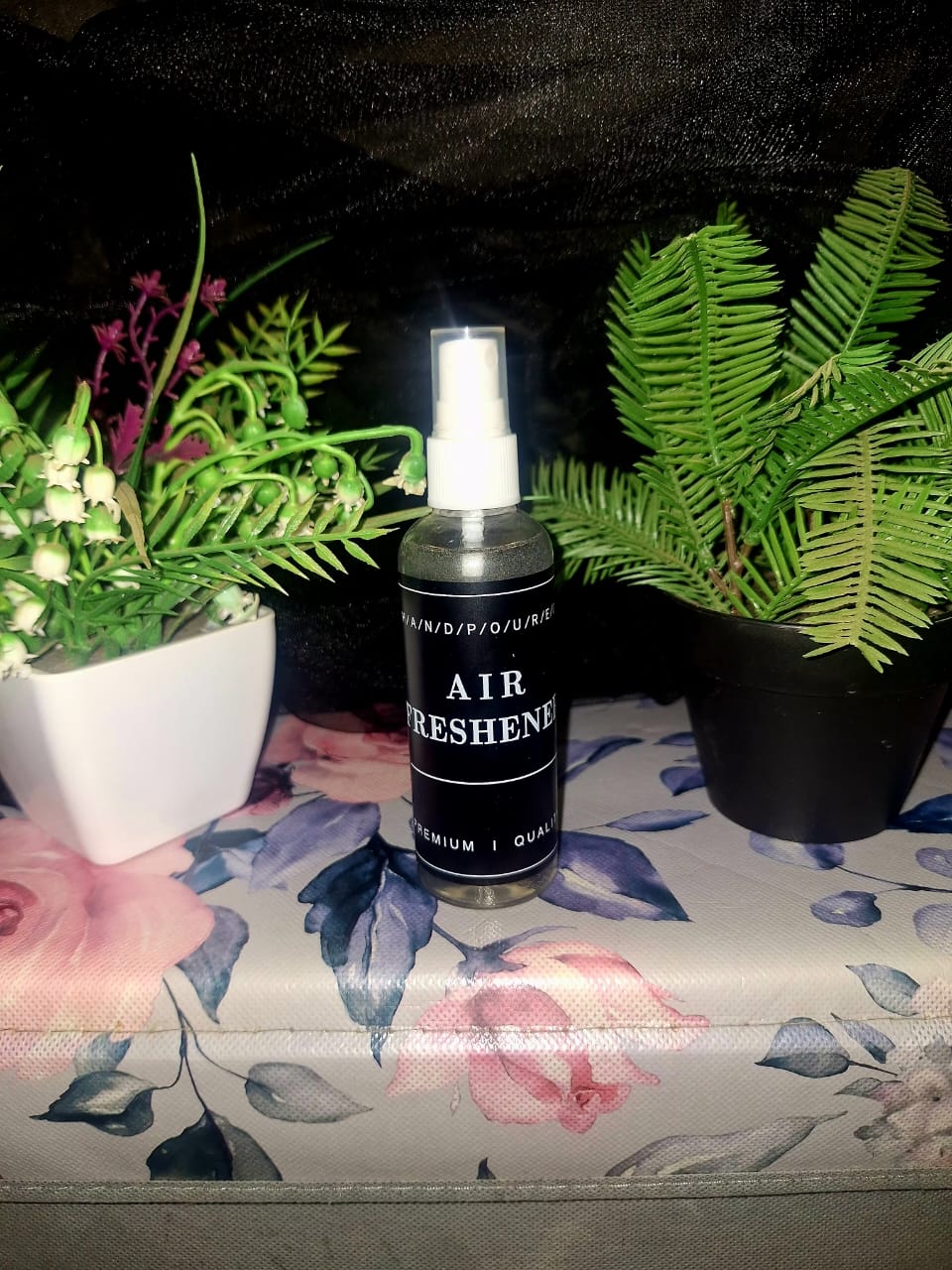 A bottle of The Dewy Petal hand-poured air freshener and linen spray, displayed on a floral surface between green botanical plants.