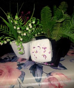 Square-shaped white botanical bath bomb with dried purple flower petals, displayed on a floral surface with green leafy plants in the background.