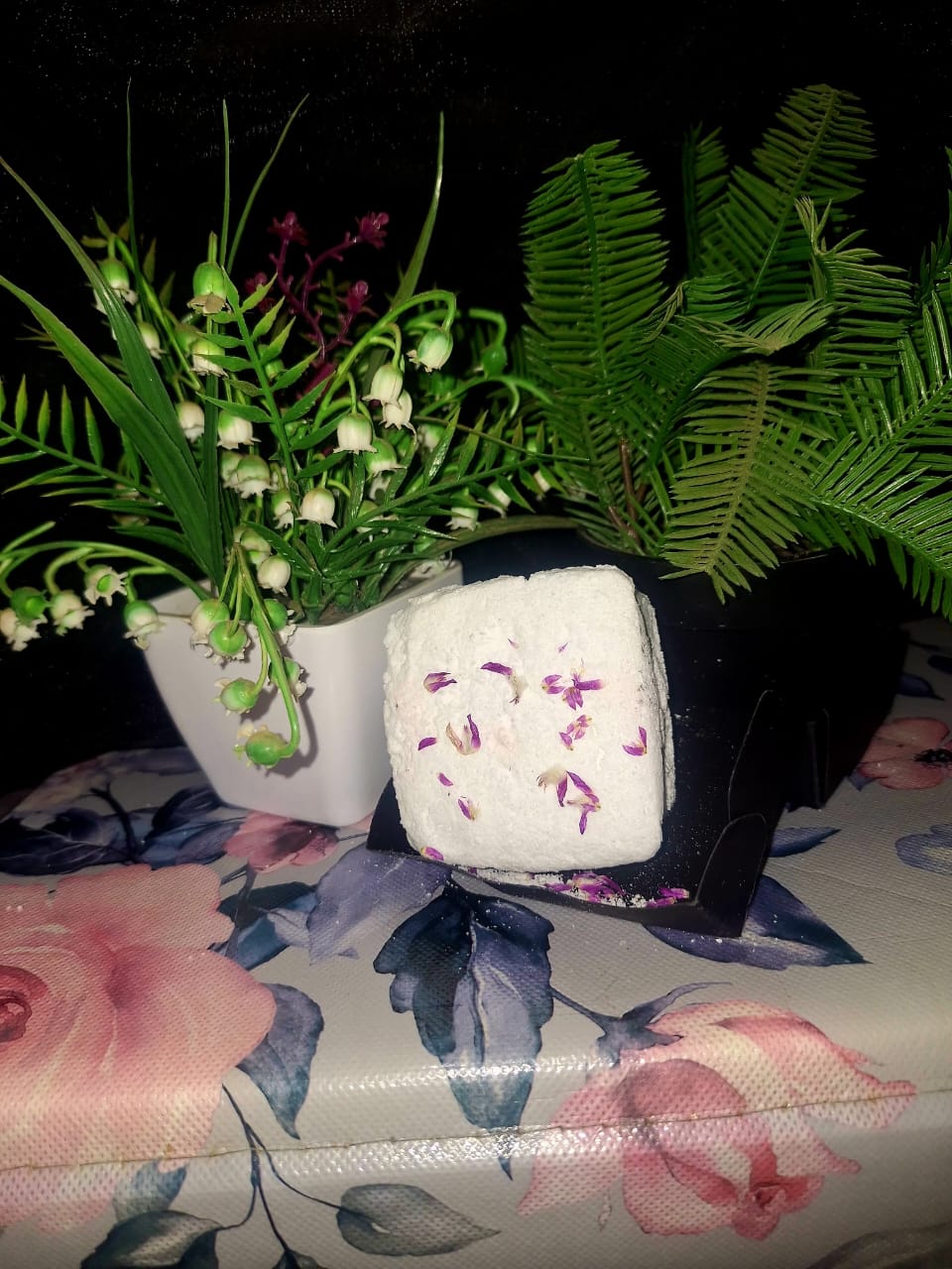Square-shaped white botanical bath bomb with dried purple flower petals, displayed on a floral surface with green leafy plants in the background.