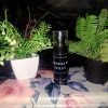 A bottle of The Dewy Petal premium essential oil spray and body mist with a clear cap, displayed on a floral rose-patterned surface between green botanical plants.