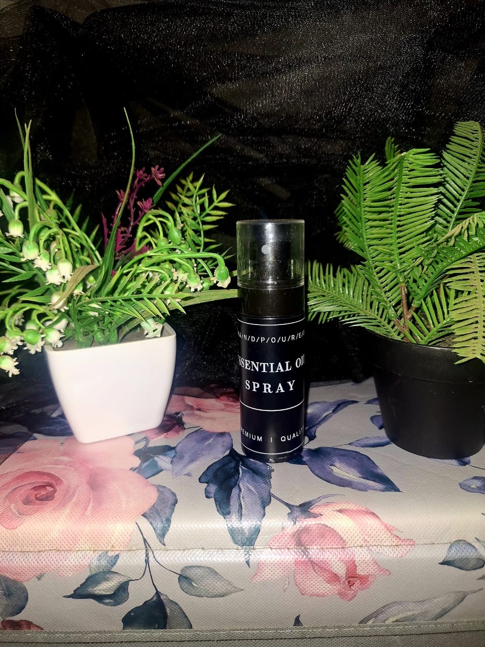 A bottle of The Dewy Petal premium essential oil spray and body mist with a clear cap, displayed on a floral rose-patterned surface between green botanical plants.