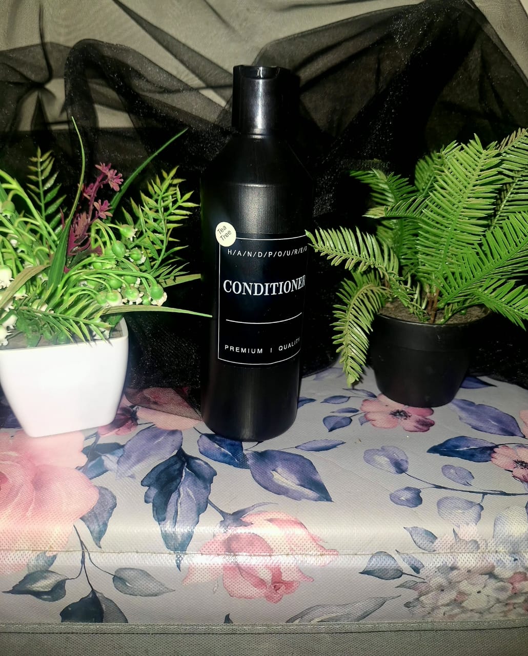 A tall matte black bottle of The Dewy Petal hand-poured Tea Tree conditioner, staged on a floral surface between green botanical plants.