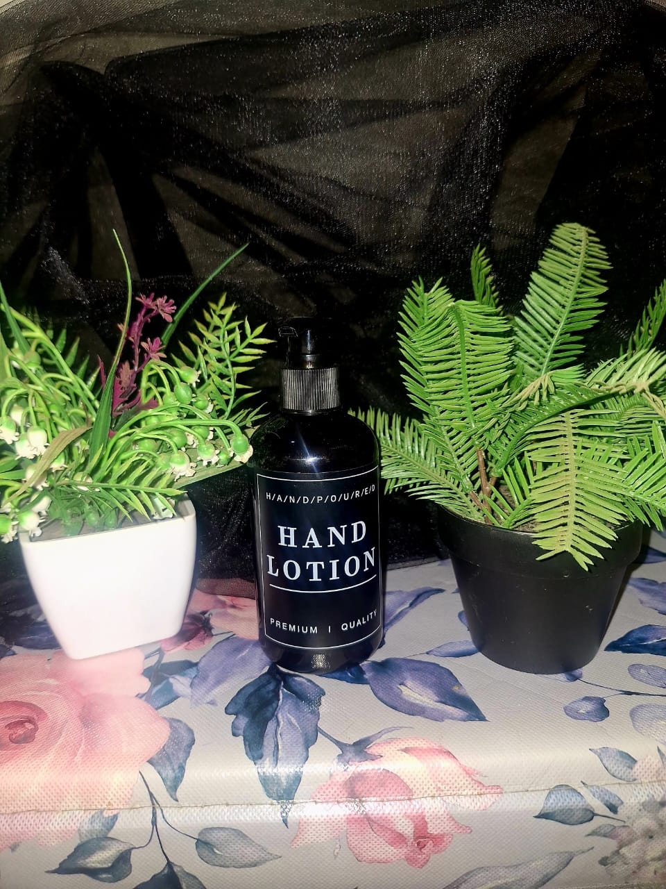 A tall black pump bottle of The Dewy Petal hand-poured hand lotion, staged on a floral rose-patterned surface between green botanical plants.