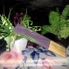 A bundle of dark purple artisanal incense sticks in clear packaging, displayed on a floral rose-patterned surface with green botanical plants in the background.