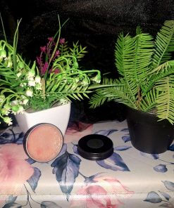 An open tin of soft pink restorative lip balm from The Dewy Petal, displayed on a floral rose-patterned surface with green botanical plants and a black lid.