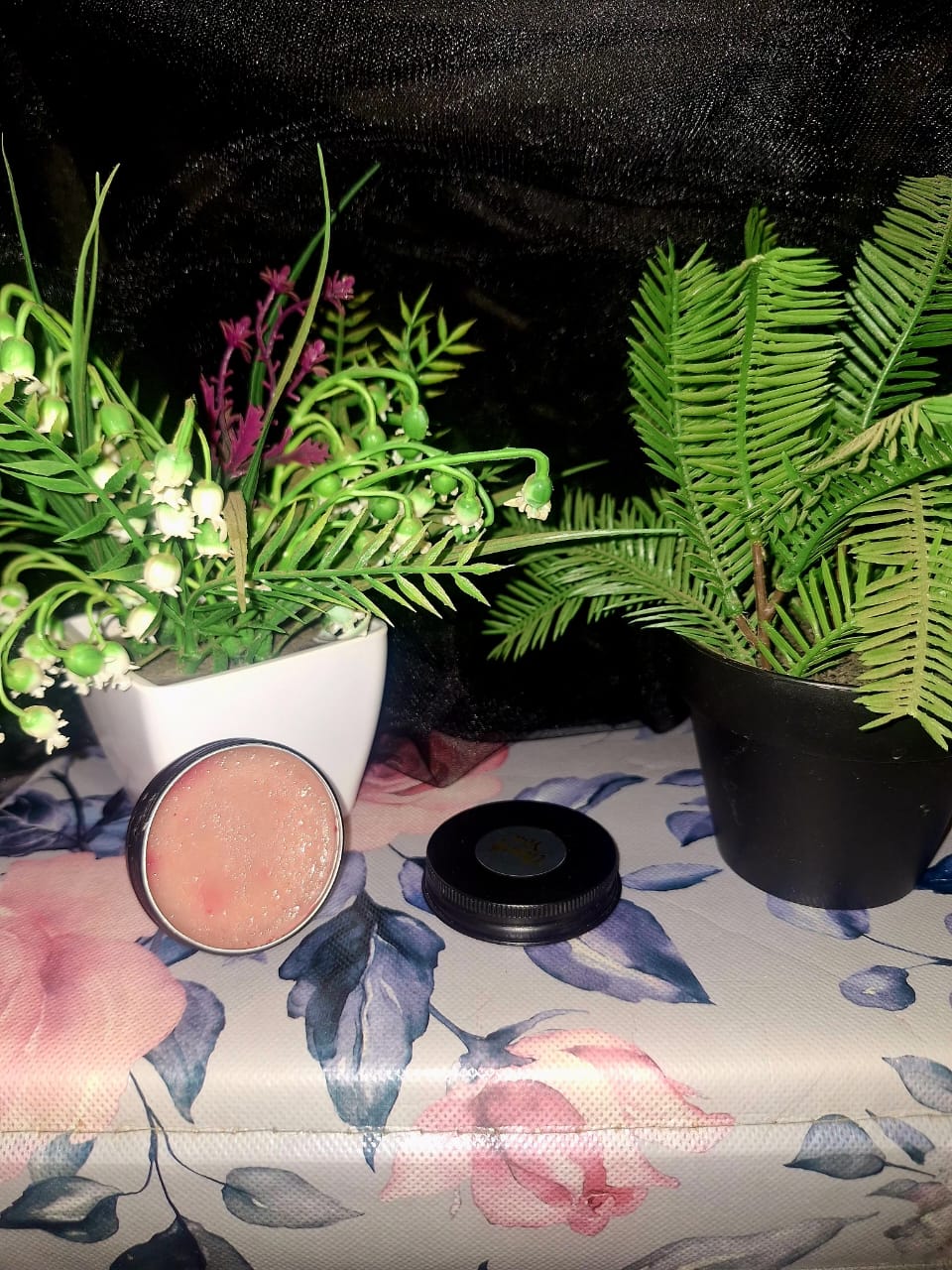 An open tin of soft pink restorative lip balm from The Dewy Petal, displayed on a floral rose-patterned surface with green botanical plants and a black lid.