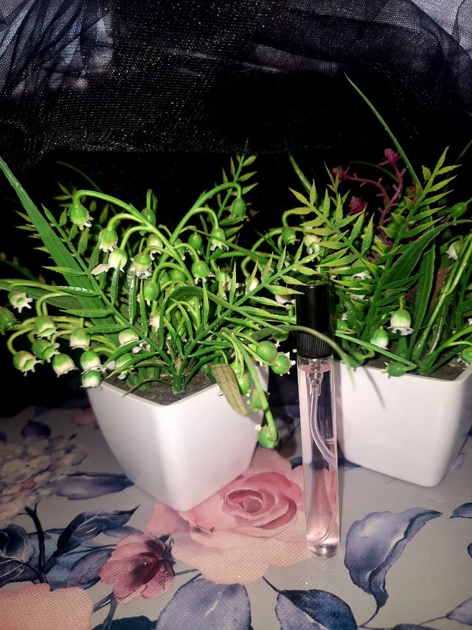 The Dewy Petal clear glass pocket spray bottle with a black cap, placed on a floral surface next to two small potted artificial plants, designed for on-the-go fragrance in South Africa.
