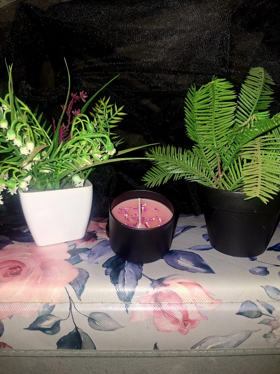 A small black tin containing a pink hand-poured candle topped with dried purple flower petals, positioned on a floral surface with botanical plants in the background.