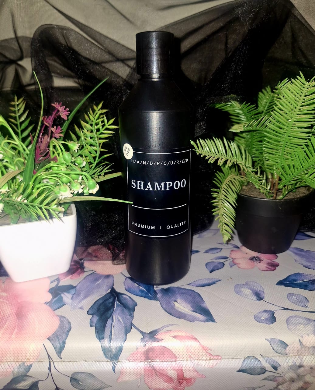 A tall matte black bottle of The Dewy Petal hand-poured Tea Tree shampoo, staged on a floral surface between green botanical plants.