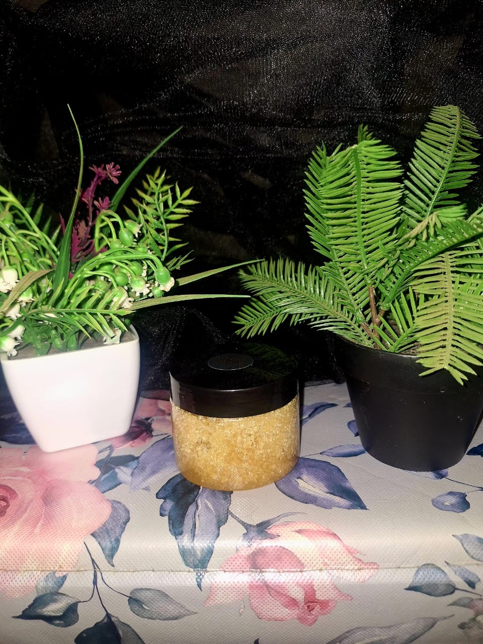 A clear jar with a black lid filled with golden-hued sugar scrub, staged on a floral rose-patterned surface between green botanical plants.