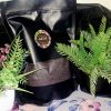 The Dewy Petal luxury bath salt in a black stand-up pouch with a floral label, displayed between green artificial plants on a floral surface.