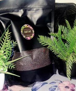The Dewy Petal luxury bath salt in a black stand-up pouch with a floral label, displayed between green artificial plants on a floral surface.