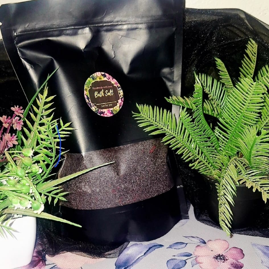 The Dewy Petal luxury bath salt in a black stand-up pouch with a floral label, displayed between green artificial plants on a floral surface.